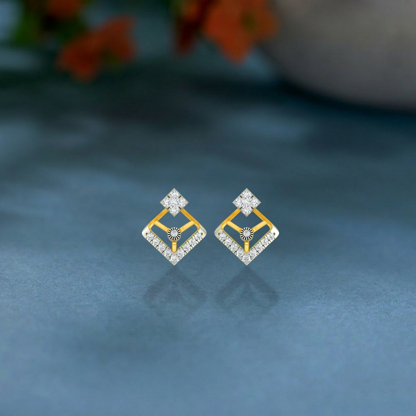 Pair of gold and diamond earrings on a dark blue surface with a blurred floral background.