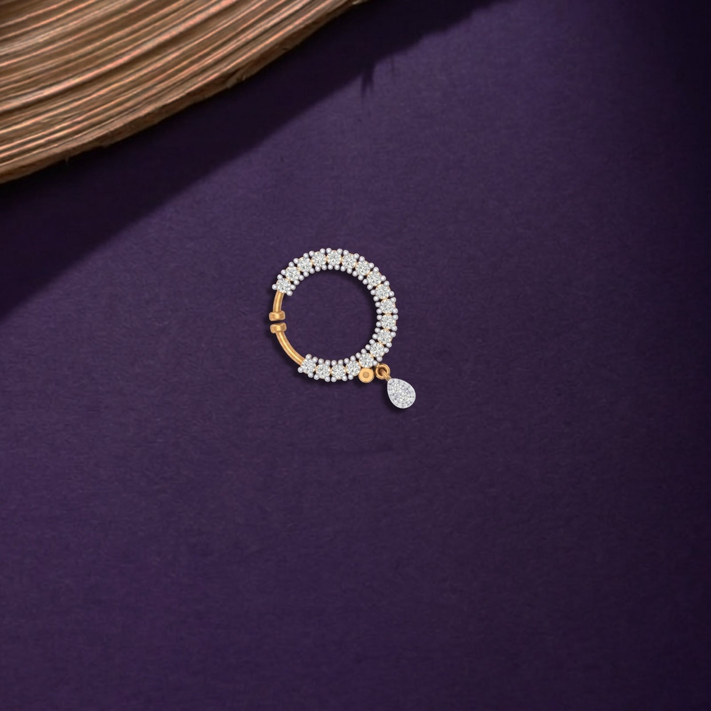 A yellow gold and diamond nose ring with a halo of small clear stones and a small teardrop-shaped clear stone pendant, displayed on a dark purple textured surface.