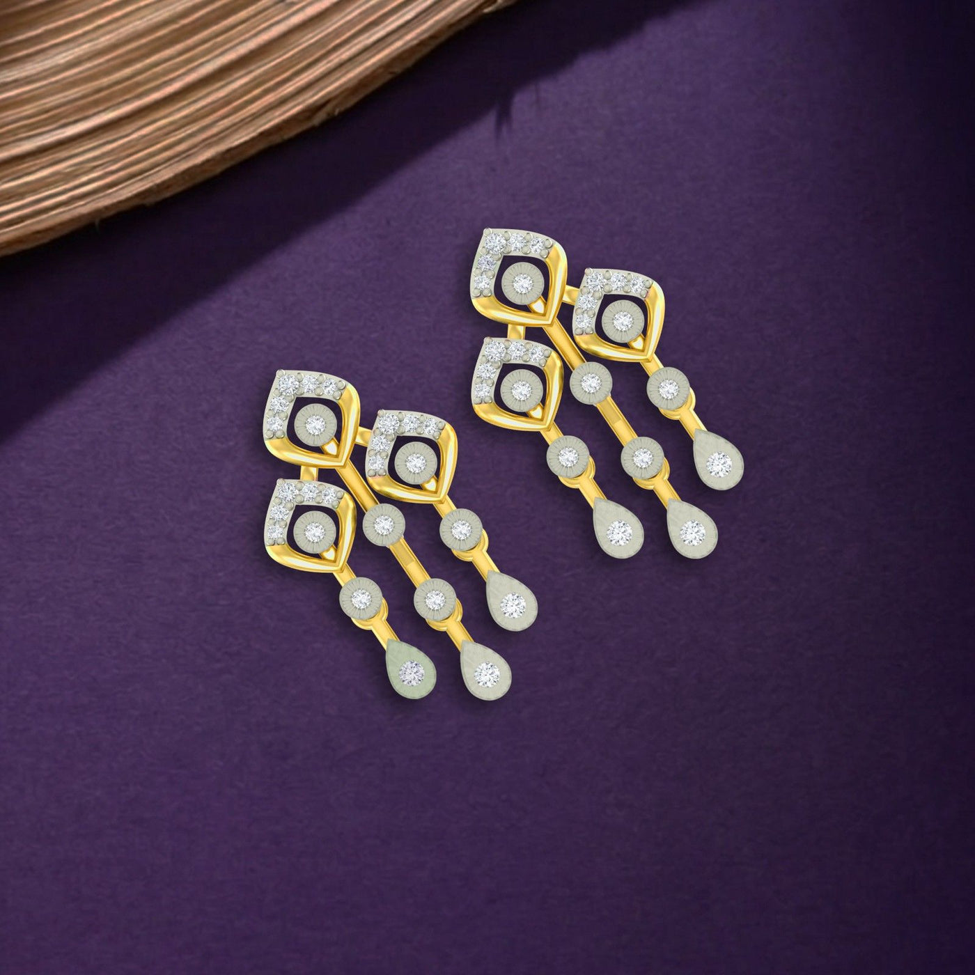 A pair of gold and diamond drop earrings displayed on a textured purple surface.