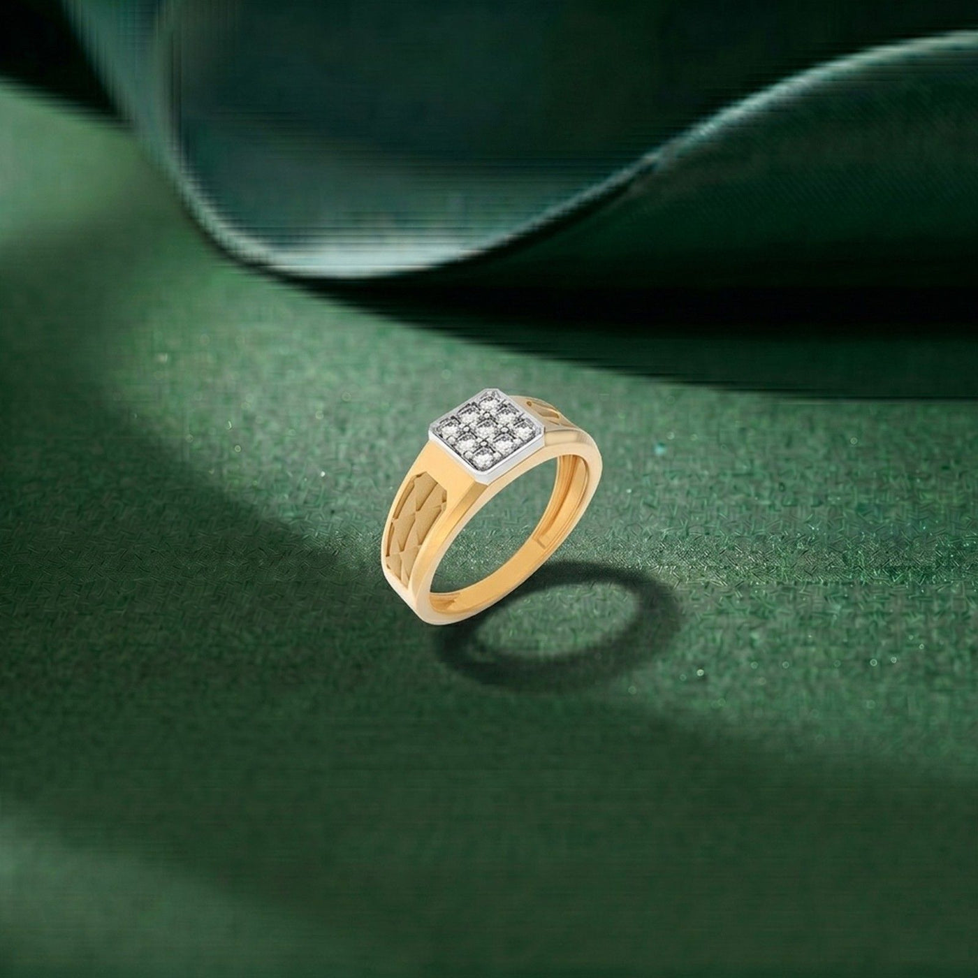 A gold ring with a square diamond cluster setting, resting on a textured dark green surface with a draped green fabric in the background.