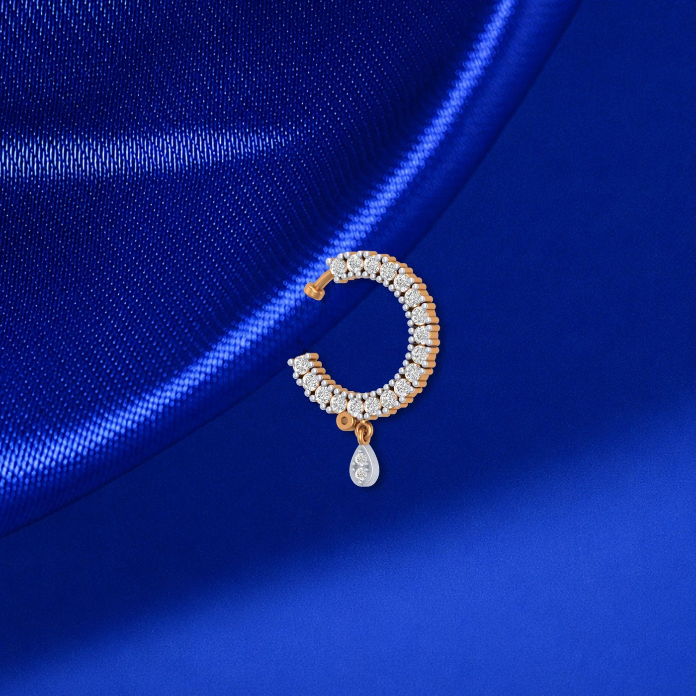 A close-up of a crescent-shaped nose ring with clear stones and a teardrop-shaped clear stone pendant, set in a warm gold-toned metal, resting on a textured royal blue satin fabric.