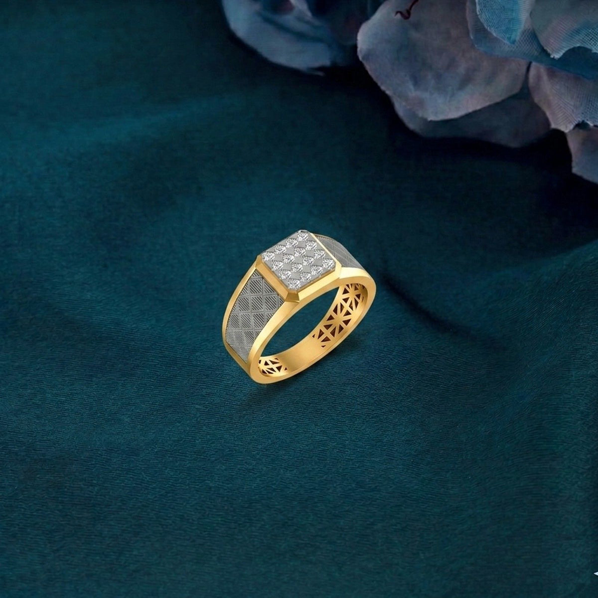 A gold ring with a square, set with small clear stones and featuring a textured geometric pattern on the band, displayed on a dark teal fabric background.